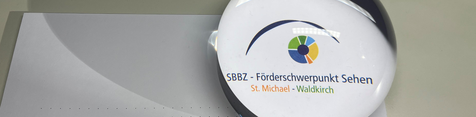 Eine runde Visolettlupe vergrößert das Logo des SBBZ.