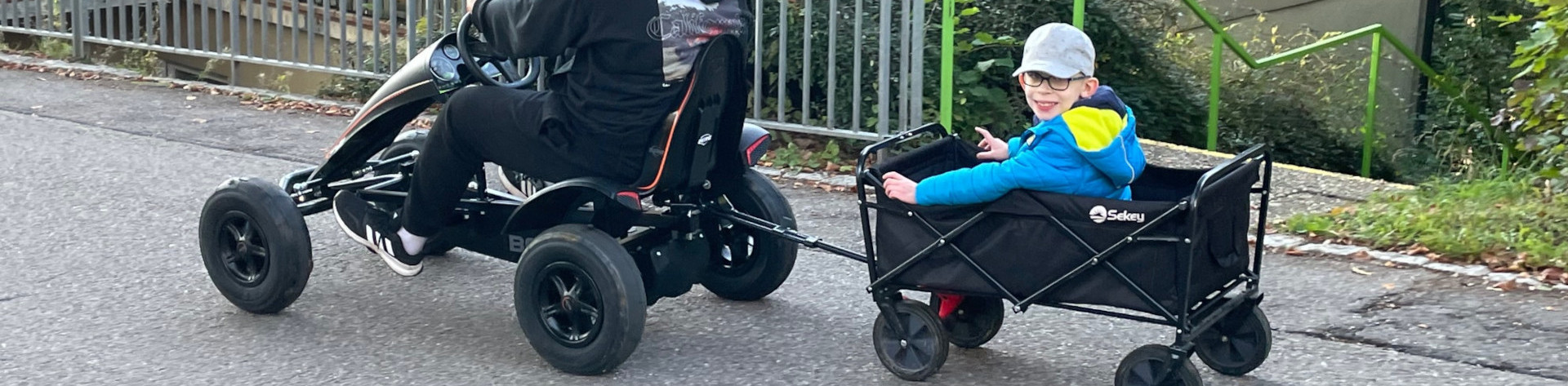 Zwei Jungen fahren auf Straße mit Go-Kart. Der größere zieht den kleineren, der in einem Anhänger sitzt und fröhlich lacht.